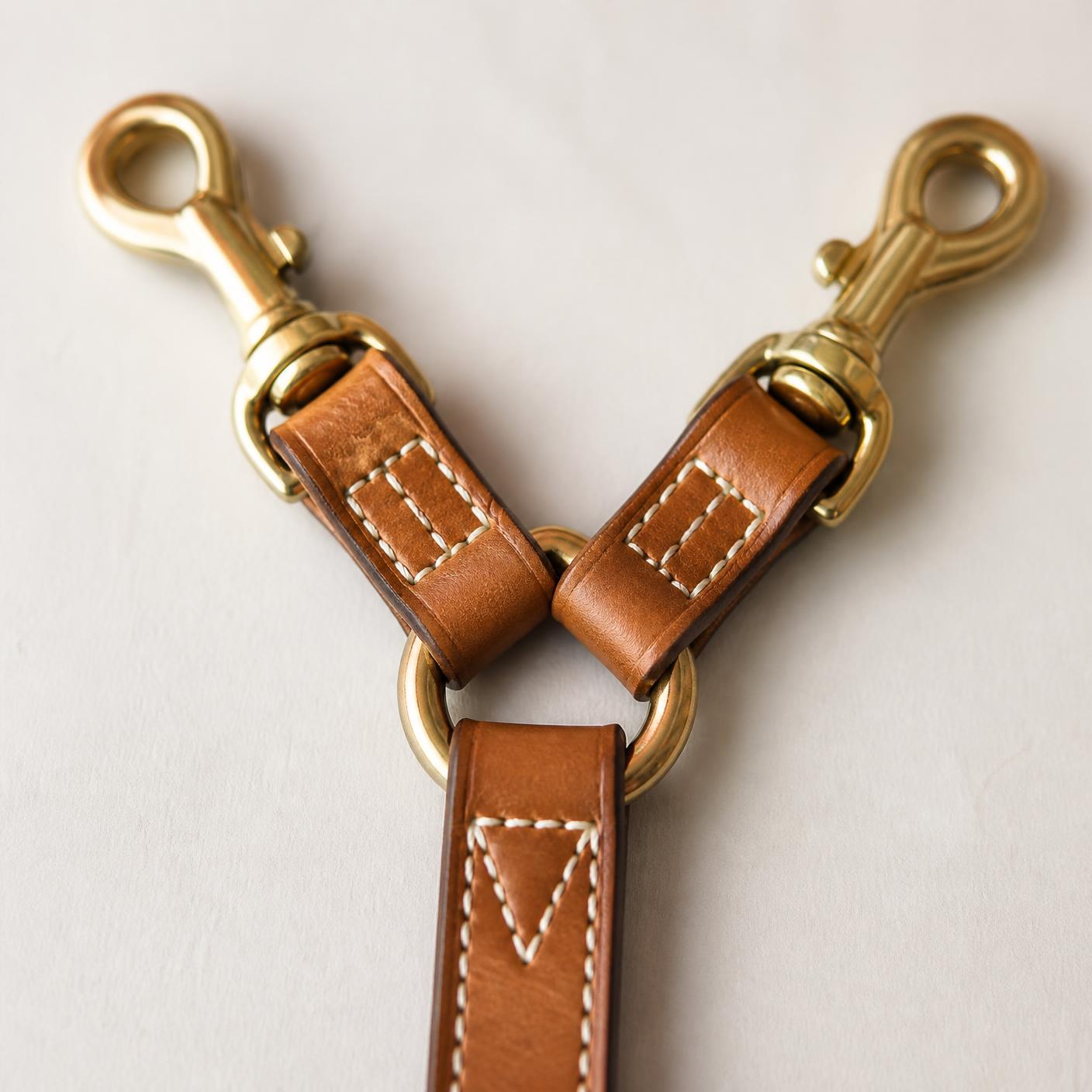 Macro detail of the leather Y junction: two hand-polished brass snap hooks, a brass ring and three saddle-stitched leather tabs.