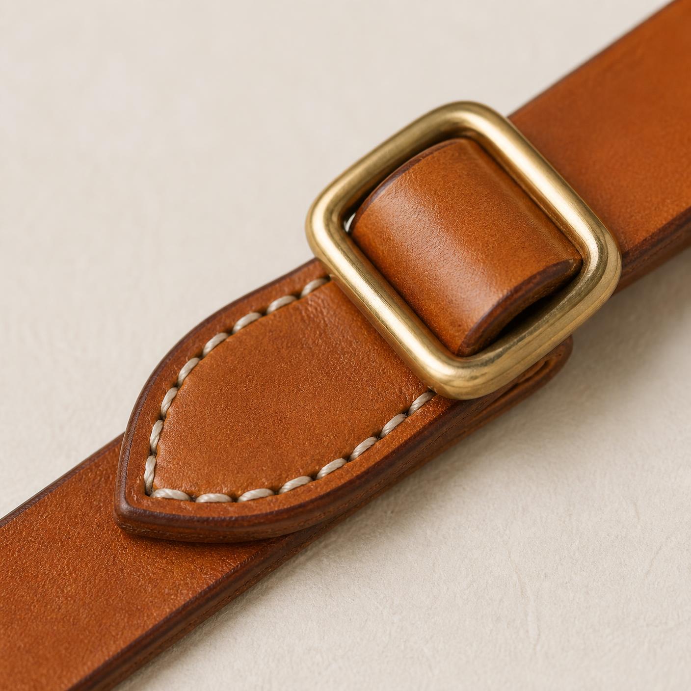 Macro detail of a brass slide buckle on natural bridle leather above a saddle-stitched leather pad.