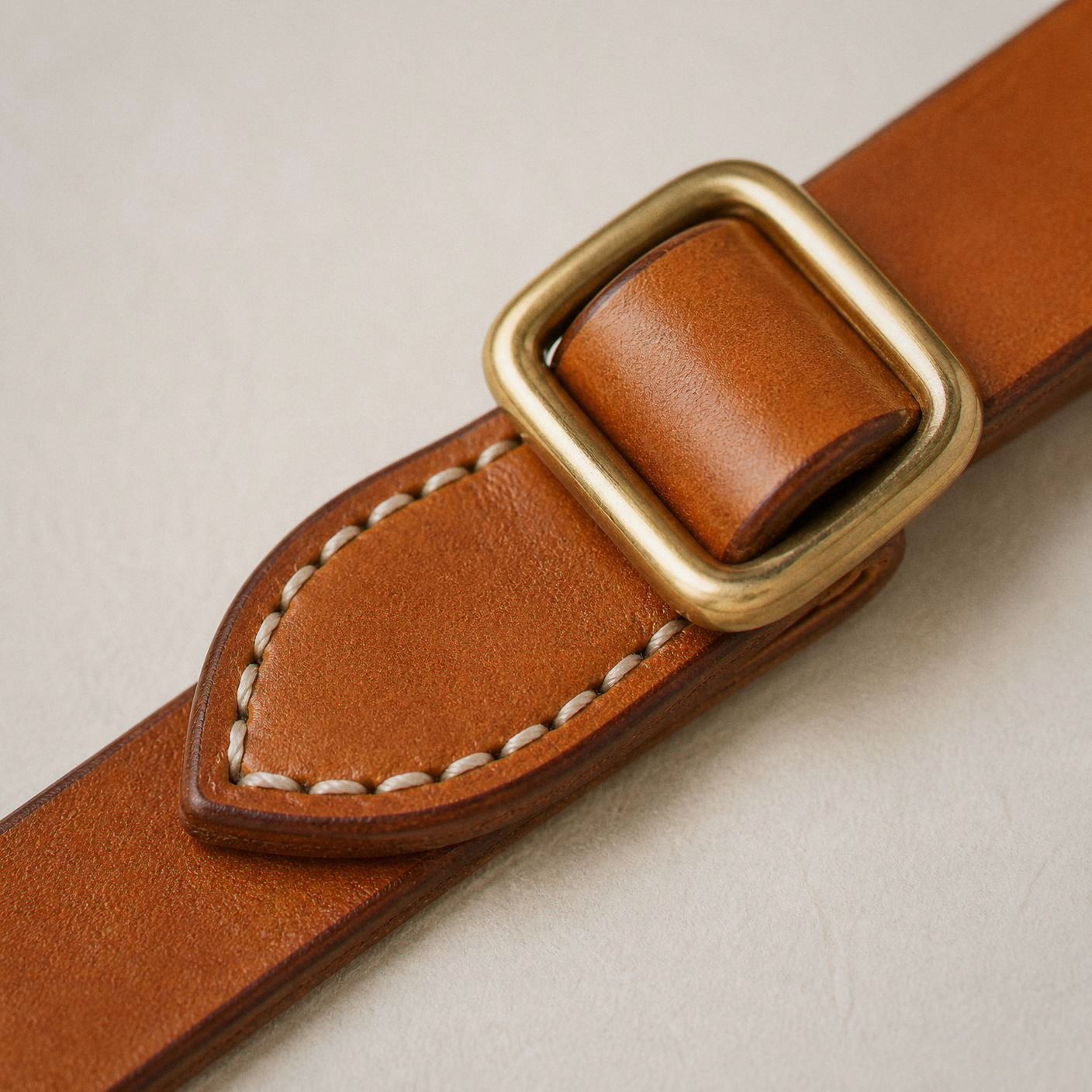 A brass slide buckle on natural bridle leather above a saddle-stitched pad.