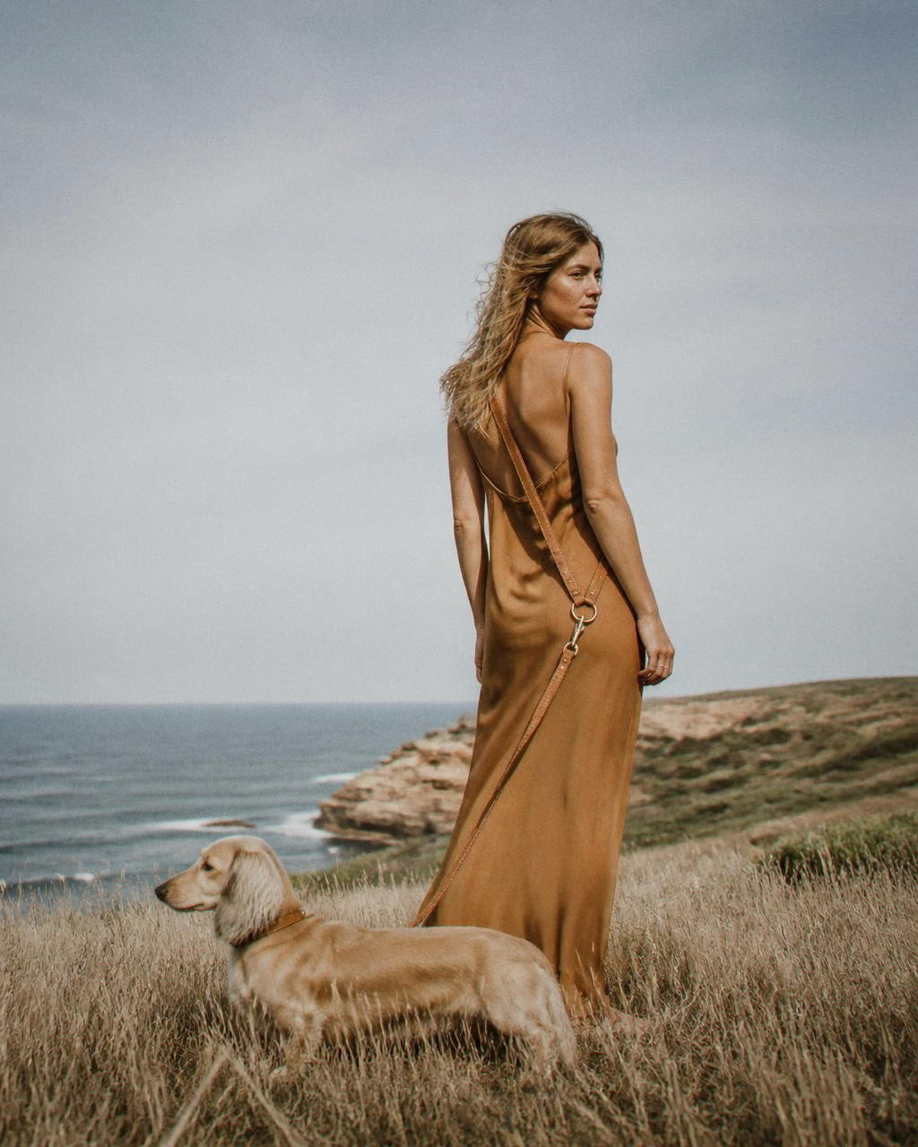 A woman stands on a dry grass headland in an ochre silk slip, the cognac leash crossbody to a cream long haired dachshund in the grass.