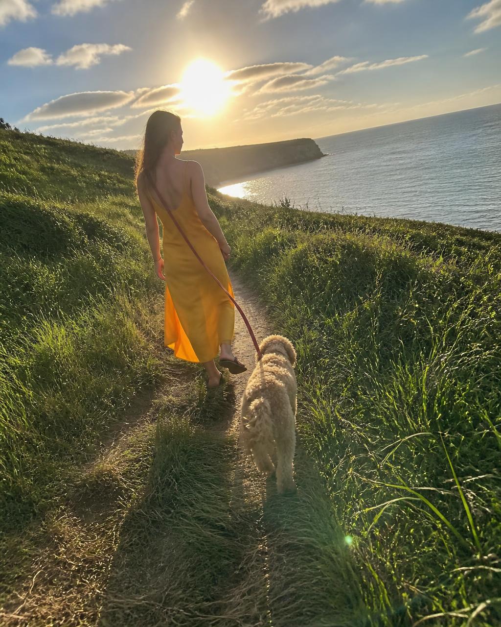 A woman walks a coastal headland at sunset in a mustard silk slip dress with her dog ahead of her.