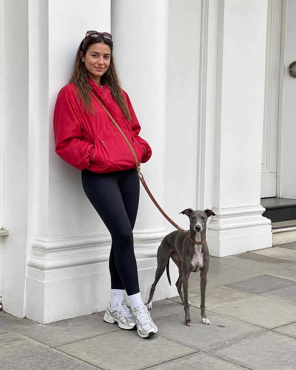 A woman in a red jacket leans against a white columned doorway with a grey whippet beside her.