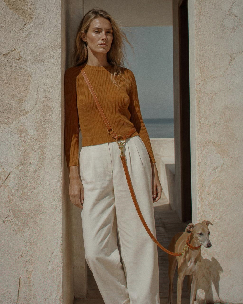 A woman leans in a limewashed doorway in an ochre knit and bone trousers with an Italian greyhound clipped to the brass ring at her hip.