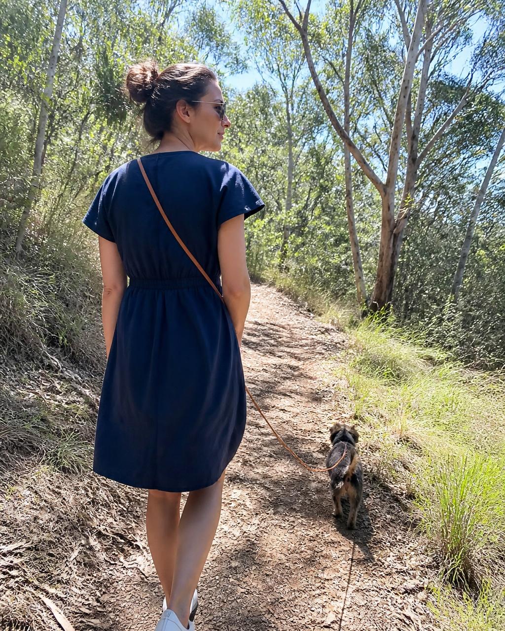 A woman walks a eucalyptus track in a navy linen dress with a small terrier ahead of her.