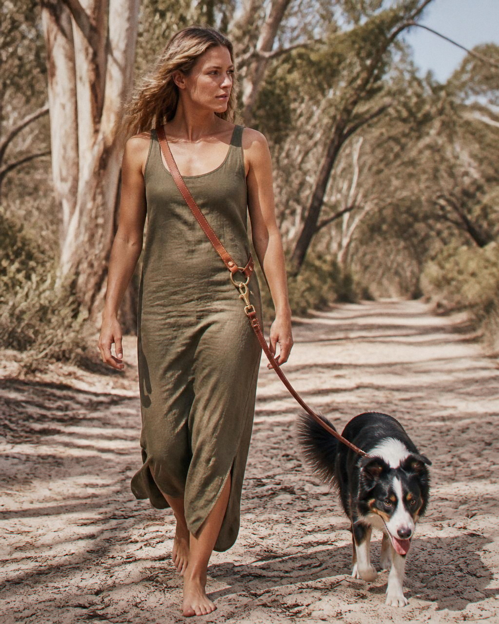 A woman walks a sandy eucalyptus track in an olive linen dress with a tricolour border collie clipped to the leash at her hip.