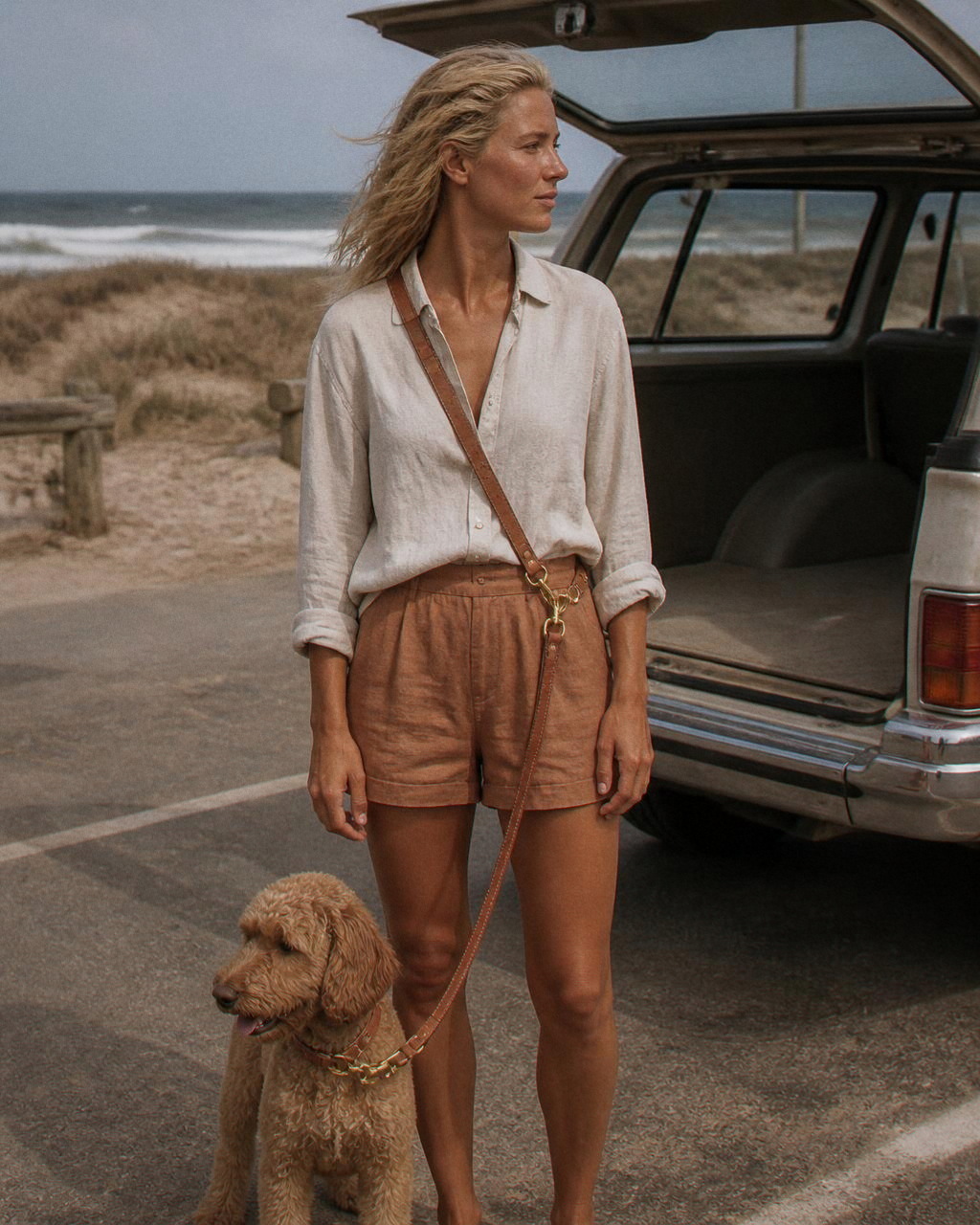A woman stands at the open tailgate of a sand coloured wagon at a beach car park with Daisy clipped to the leash at her hip.