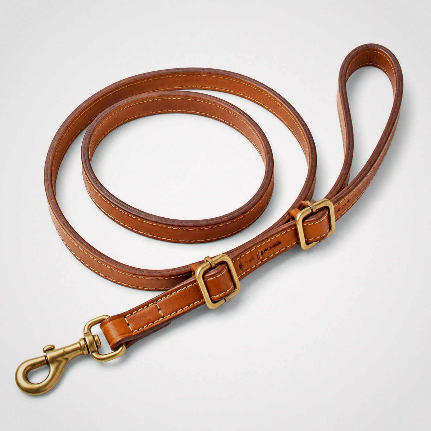 The Boundless Leash laid flat: a tan natural bridle leather strap coiled loosely with two brass slide buckles and a brass swivel clip.