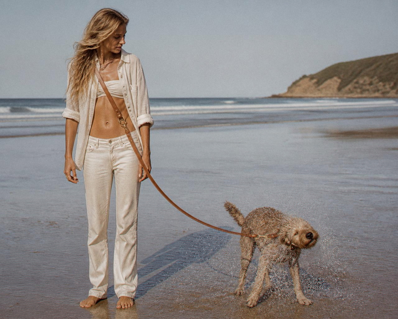 Daisy shakes seawater from her coat on flat wet sand, hands empty beside her, the lead worn crossbody.