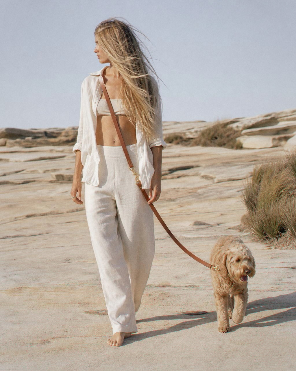 The Boundless leash in cognac saddle-tan leather worn crossbody from shoulder to opposite hip, closed at the hip with a polished brass trigger snap, the lead running taut to a dark blonde spoodle walking ahead.