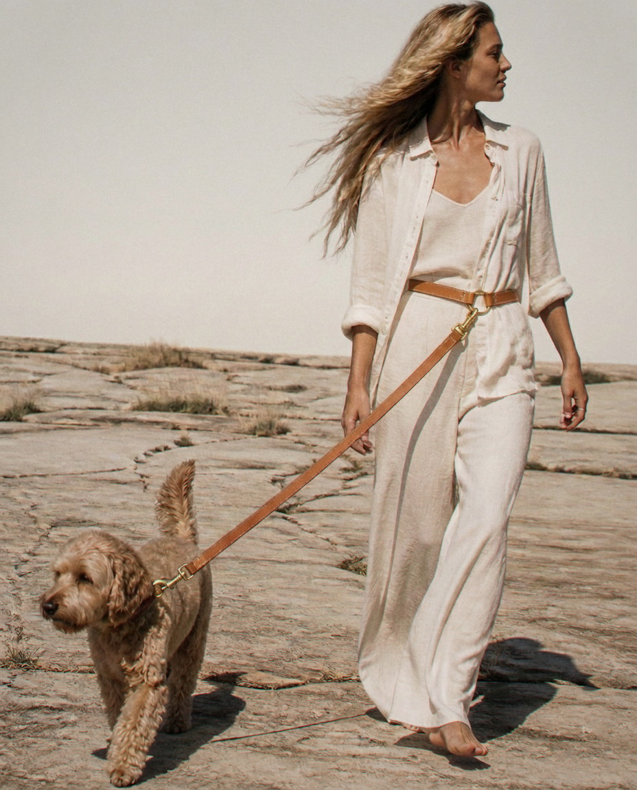 The Boundless leash worn around the waist over bone-pale linen, closed at the front hip through a brass ring, the lead running taut forward to a dark blonde spoodle walking ahead.